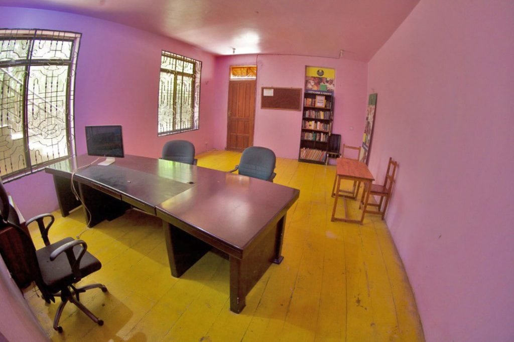 Pink study room with a dark large desk, computer monitor, and two blue office chairs on a yellow wooden floor; windows with bars and a bookshelf in the corner.