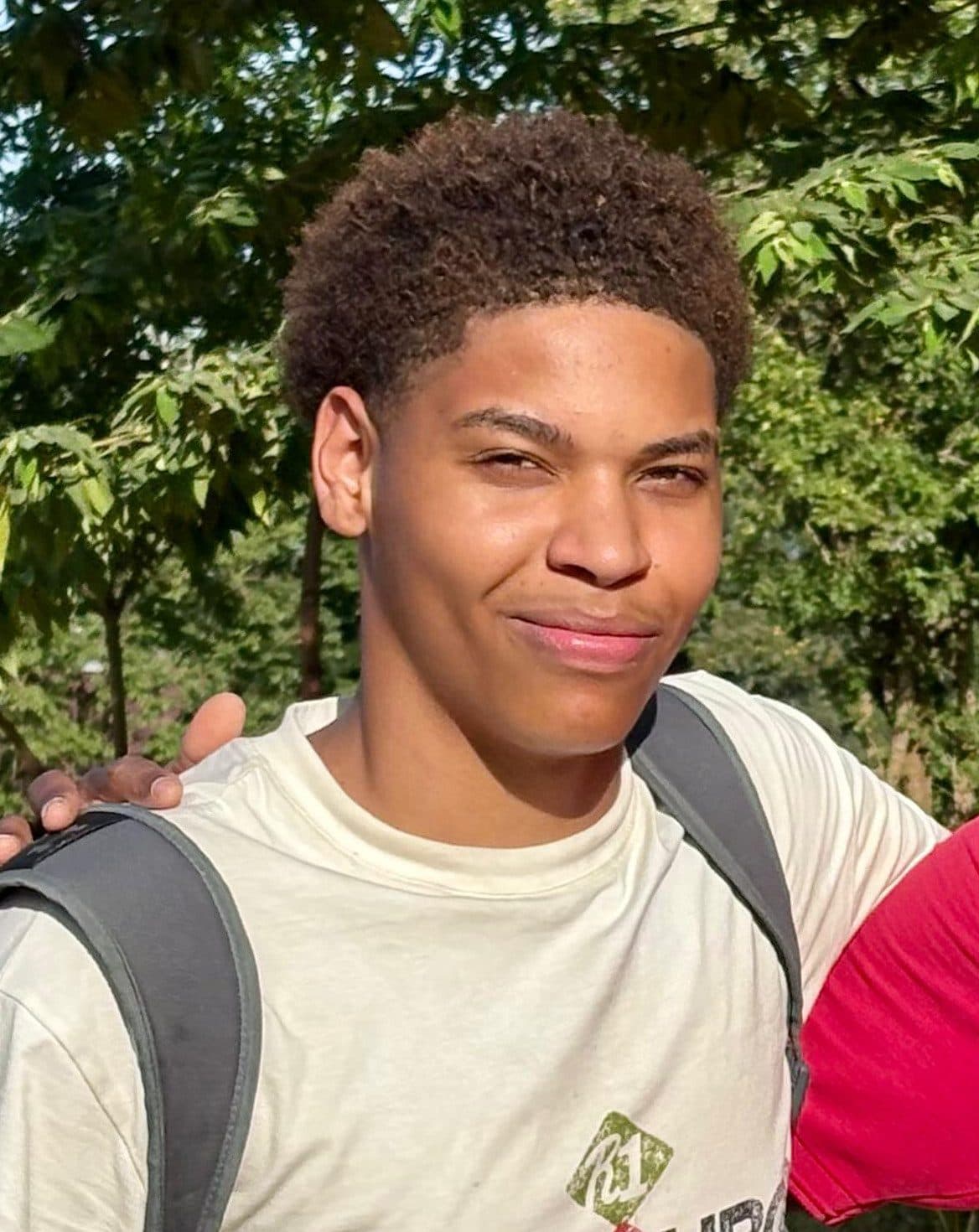 Smiling young man outdoors, wearing a light shirt and a backpack, with greenery in the background.