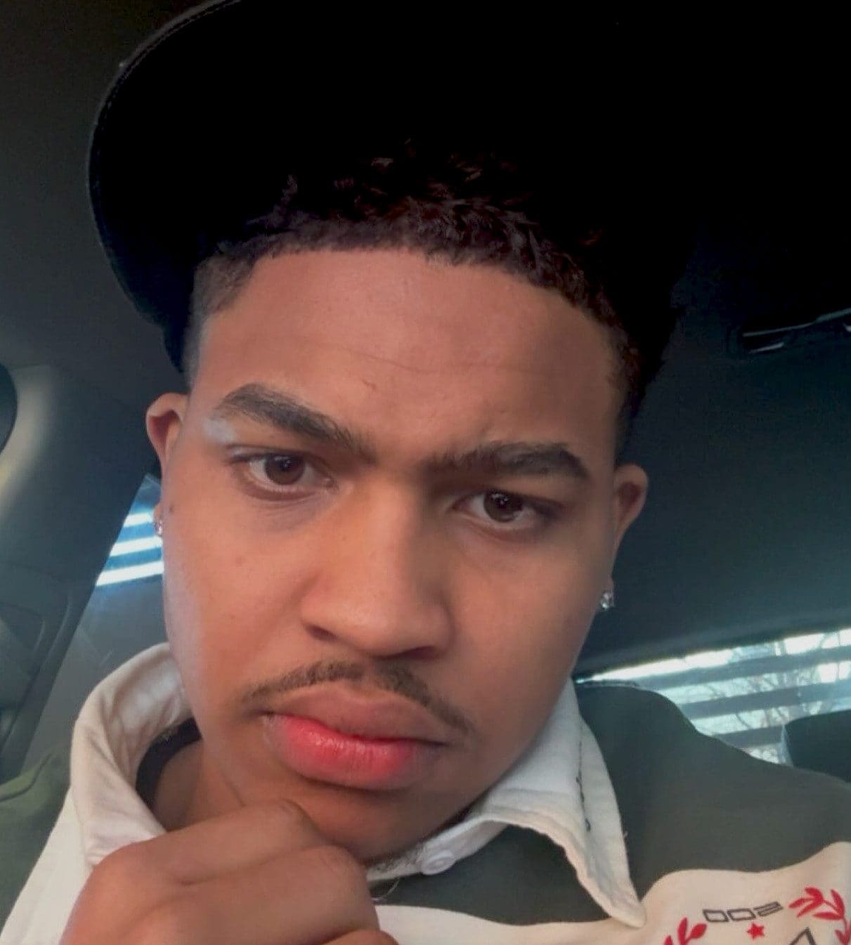 Close-up of a young man wearing a black cap and white collared shirt, resting his chin on his hand inside a car, with a serious expression and an earring visible.