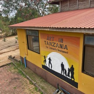 Yellow building with a mural that reads 'Art in Tanzania for Community Development' and silhouettes of people against a large white sun at sunset on the wall, outdoors in a rural area.