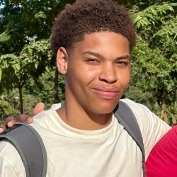 Huston Smiling young man outdoors, wearing a light shirt and a backpack, with greenery in the background.