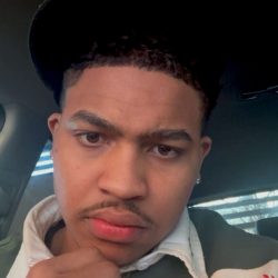 Kimi Close-up of a young man wearing a black cap and white collared shirt, resting his chin on his hand inside a car, with a serious expression and an earring visible.